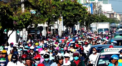 A street is full and crowded with drivers on motor bikes and cars, most wear masks and helmets.