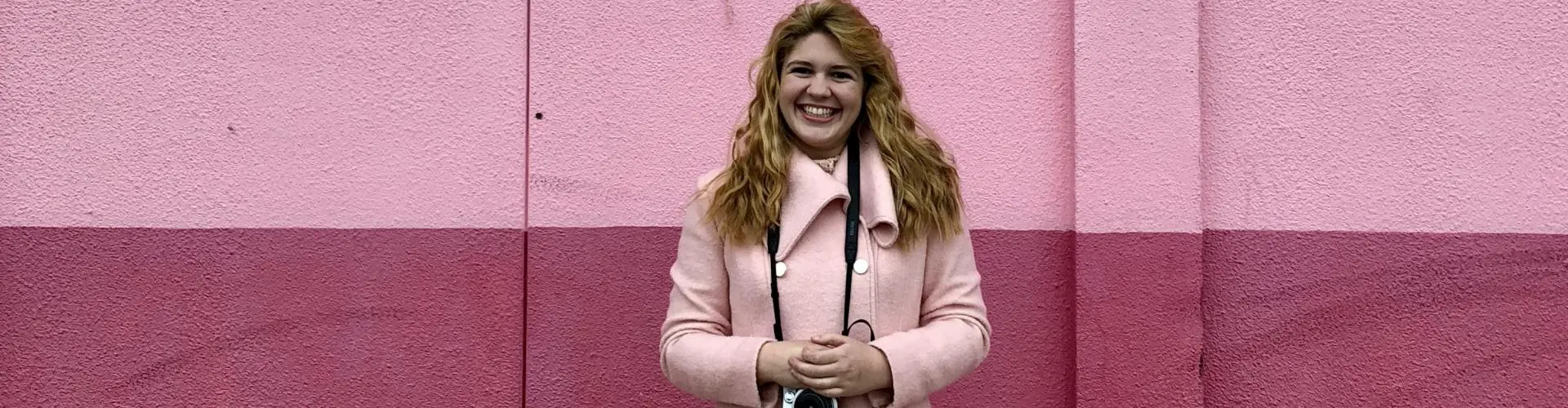 Student in pink jacket in front of pink wall with a camera.