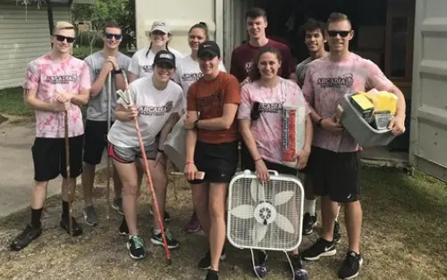 10 students, most of whom wear Arcadia t-shirts, stand together holding a large fan, a box of books, and gardening hoes