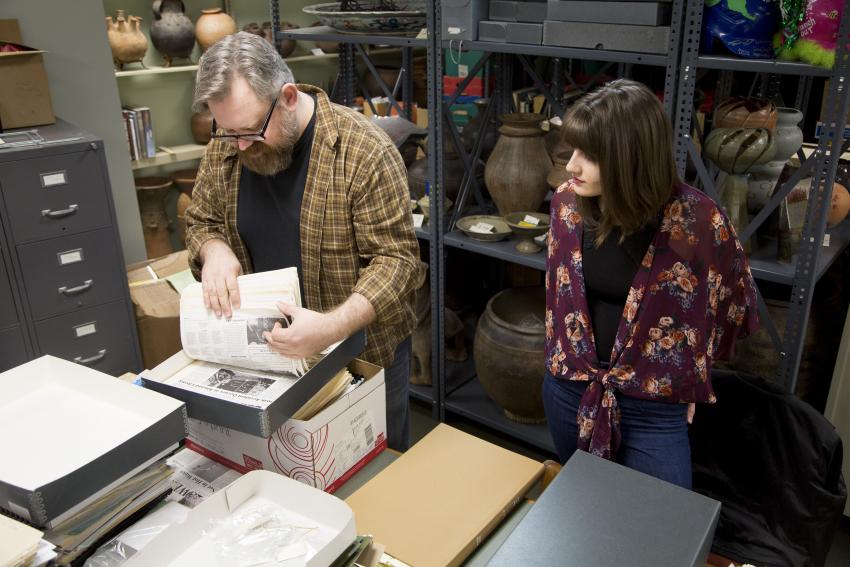 A man and woman going through old newspapers on a desk with old pottery on a shelf behind them