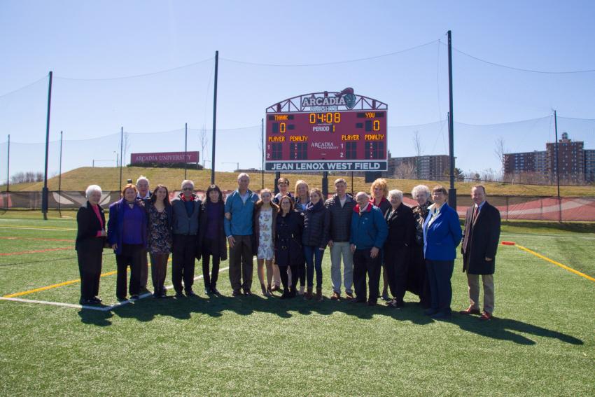 A large group wearing coats stand in front of the scoreboard and sign for Jean Lenox West Field on a sunny day.