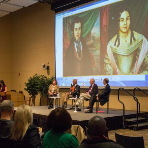 A group of speakers with two paintings projected behind them in the Great Room