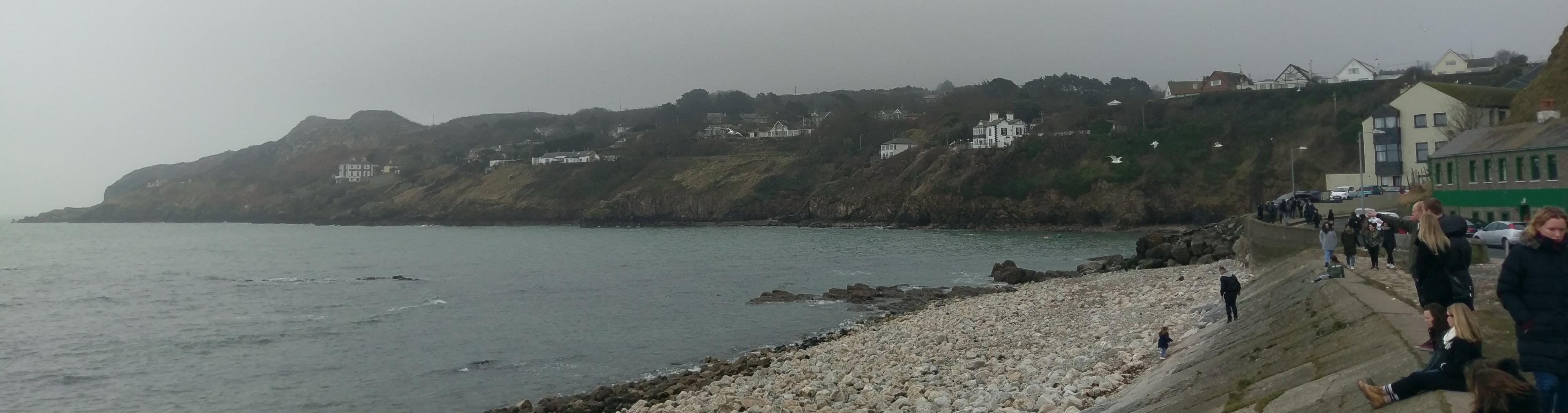 A landscape of a beach that is rocky in the foreground and hilly with sporadic homes in the background