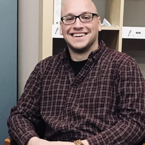 A man wearing glasses and a purple plaid shirt smiles at the camera with a laptop and white cup on the table beside him and cubbies behind him