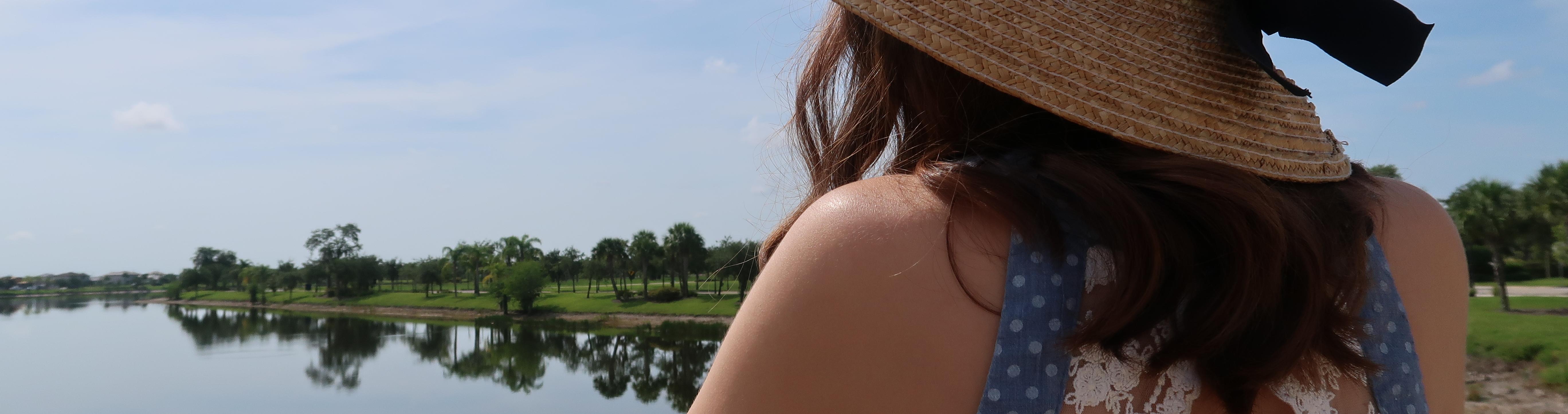 A woman's back is turned to the camera as she gazes at a body of water reflecting the trees beyond