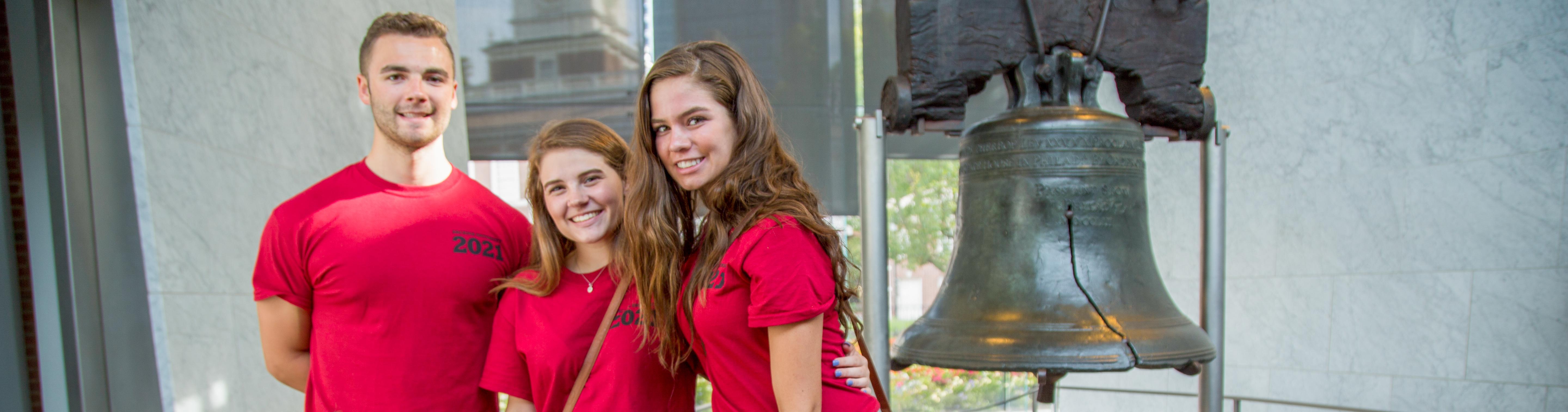 Three students wearing red shirts pose in front of the Liberty Bell