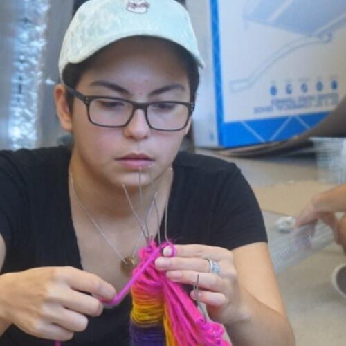 Student knitting in class while a background student works on a project.