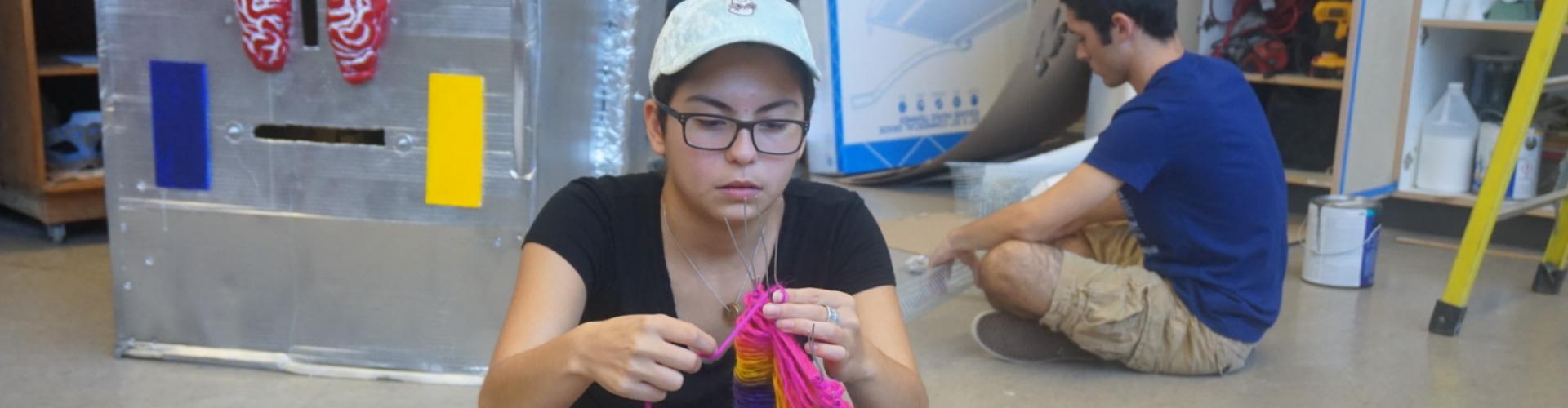 Student knitting in class while a background student works on a project.