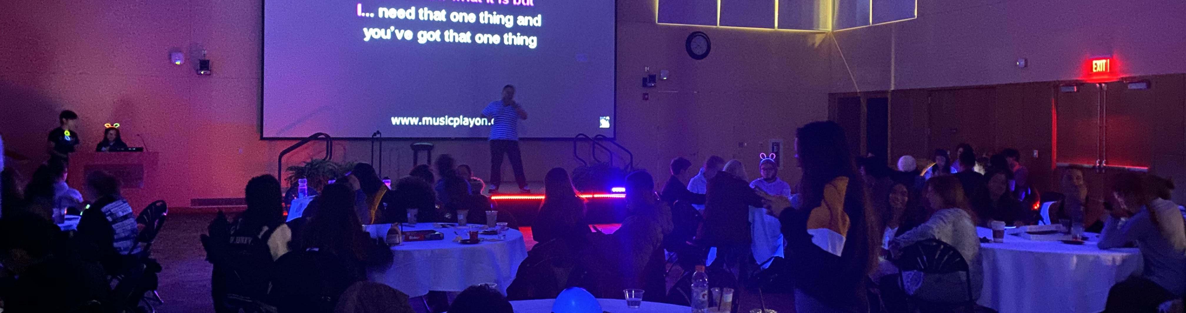 A man on stage in the Great Room with a crowd.