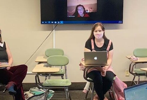 Masked students sit in a circle of desks with a large screen and zoom attendee behind