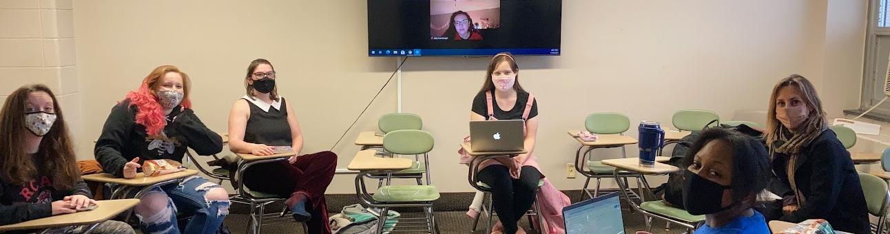 Masked students sit in a circle of desks with a large screen and zoom attendee behind