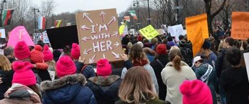 Sign that says "I'm with Her" pointing towards the crowd surrounding it in a street.