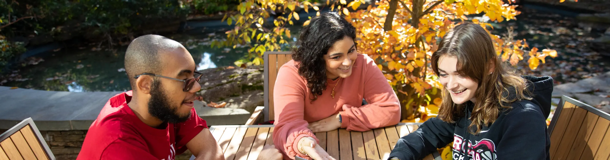 Three students sit together at a table outside of Commons with yellow fall leaves on the tree behind them