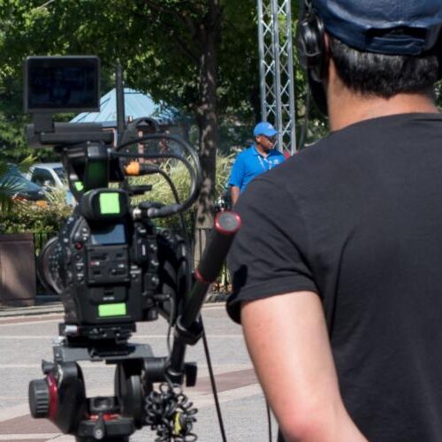 A young woman with blonde hair being filmed by two men as she talks while wearing a dark blue "ACEing Autism" shirt.