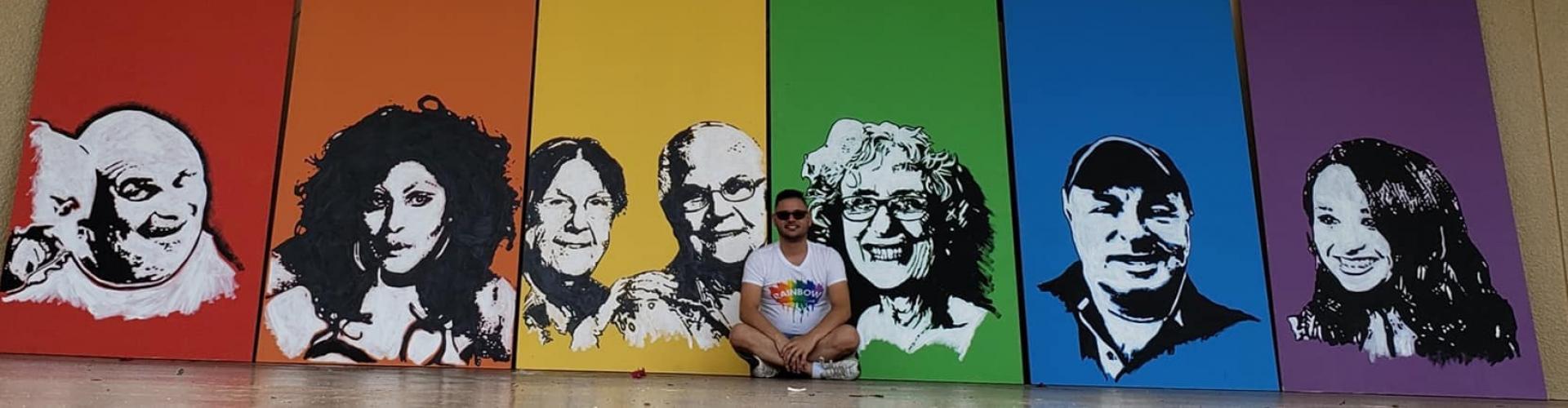 A student sits in front of a rainbow colored mural