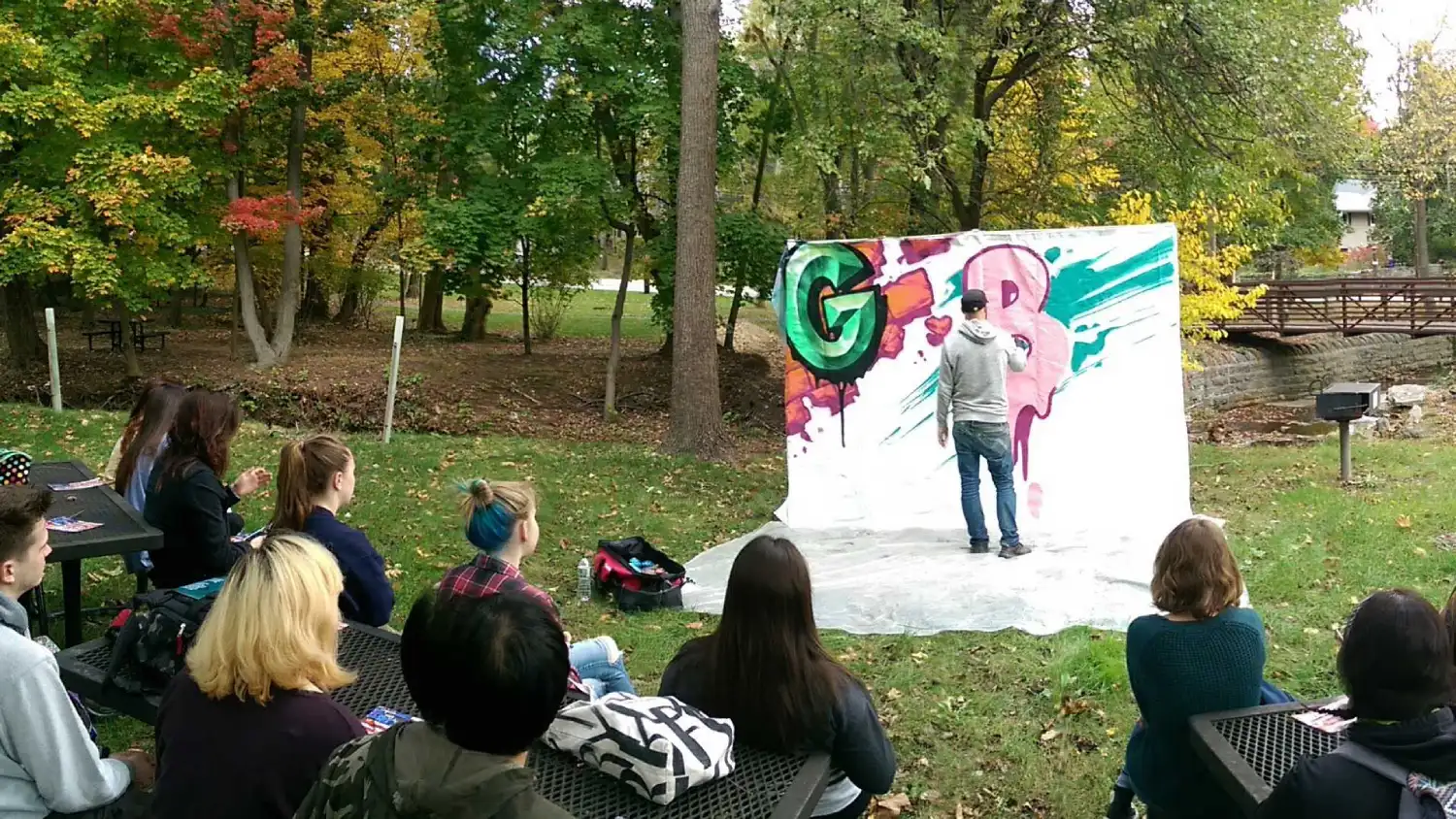 Group of students watch artist spray paint artwork onto white background in park.