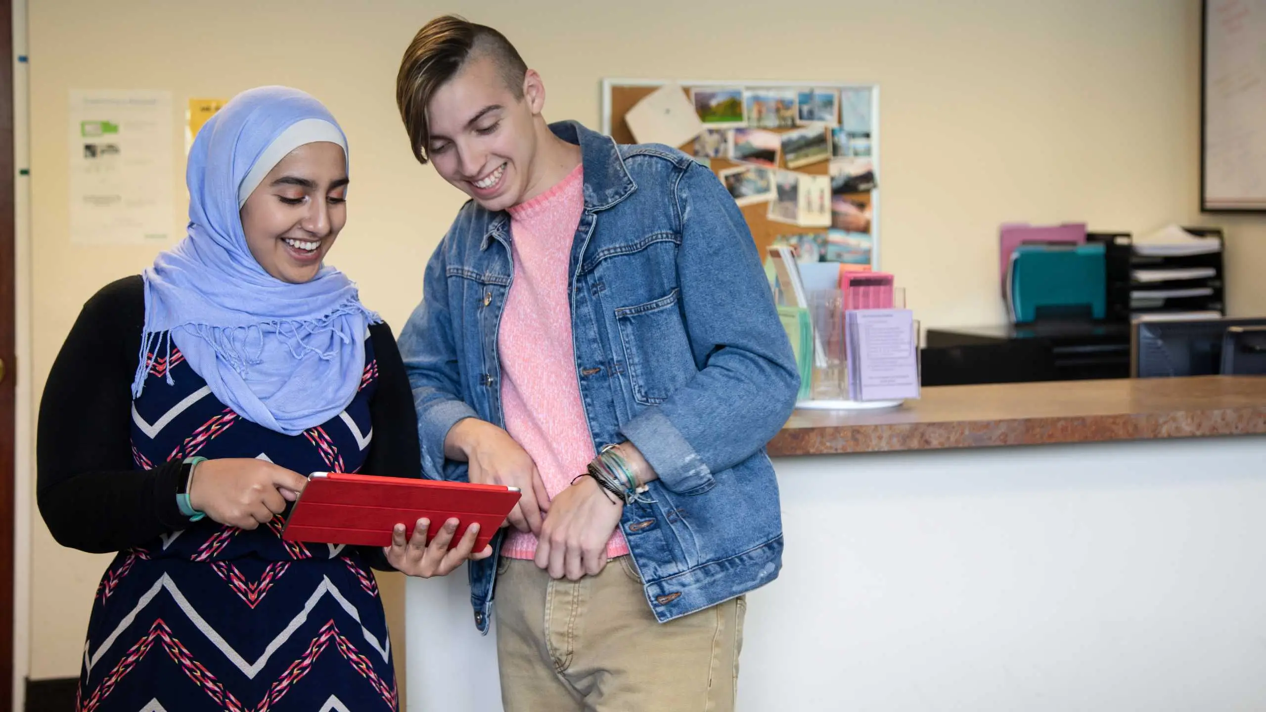 Two students standing while leaning on a counter, smiling at a tablet device