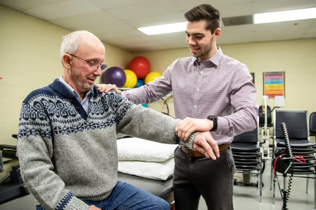 A physical therapy student works with an older male patient who is seated.