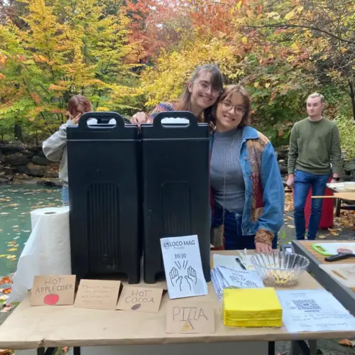 Two students smiling at hot cider, cocoa, and pizza stand.