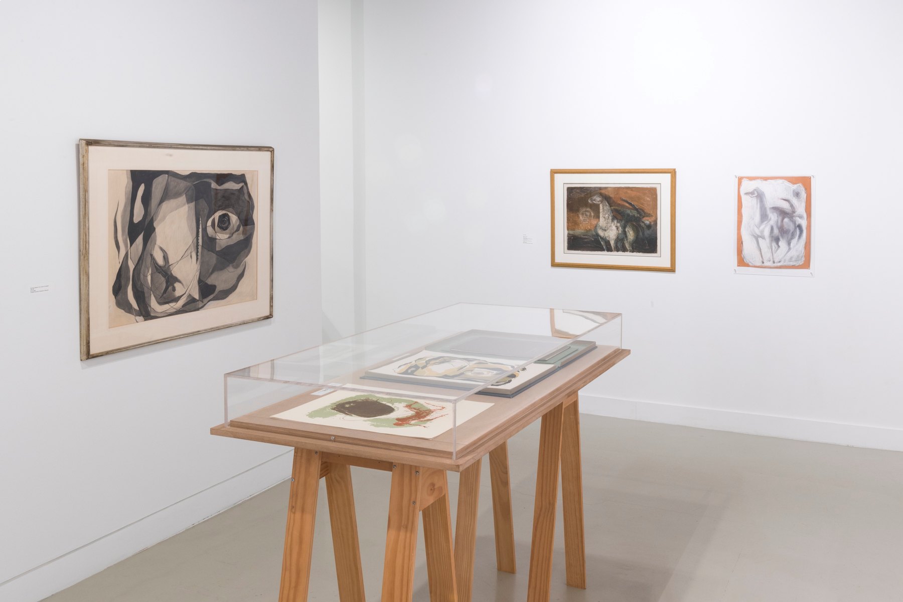 A wooden legged glass display case exhibiting artwork in the Spruance Gallery with other pieces of artwork on the walls