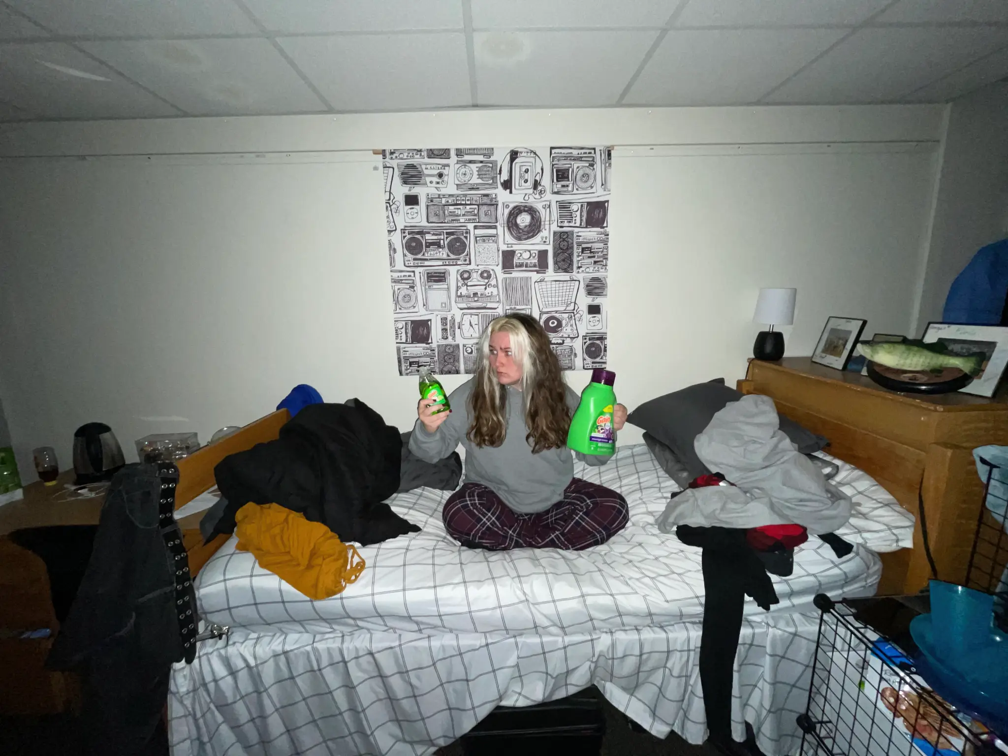 A student sits on a form room bed holding dish soap in one hand and laundry detergent in the other