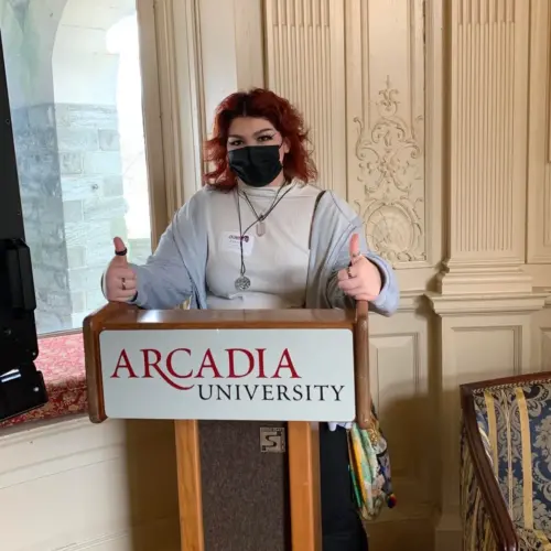 A student wearing a face mask stands at an Arcadia University podium and gives two thumbs up