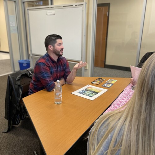 Alum Tim Smith sits at a table with artwork in front of him talking to students