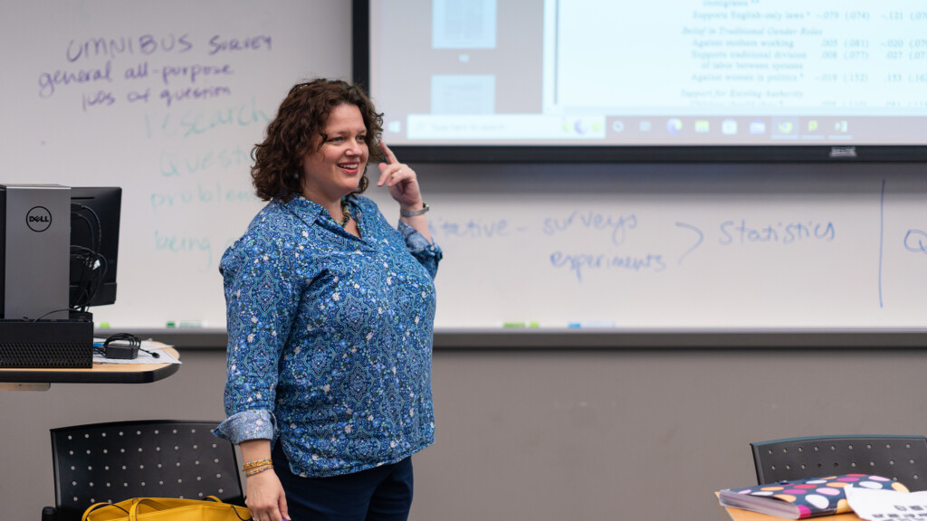 Dina Pinsky teaches and stand in front of a screen smiling.