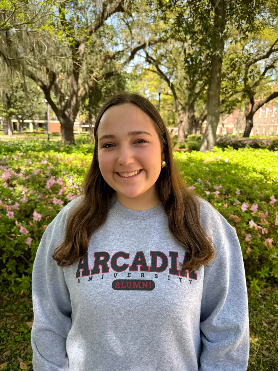 Young woman with brown hair in a grey Arcadia sweatshirt. Standing in front of a tree.