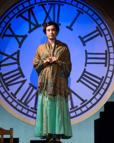 A theater performer wearing a shawl and turquoise shirt standing on podium with large clock behind them