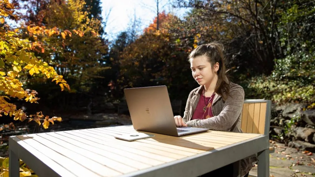 A student sitting outside during the day at a table, working on a laptop.