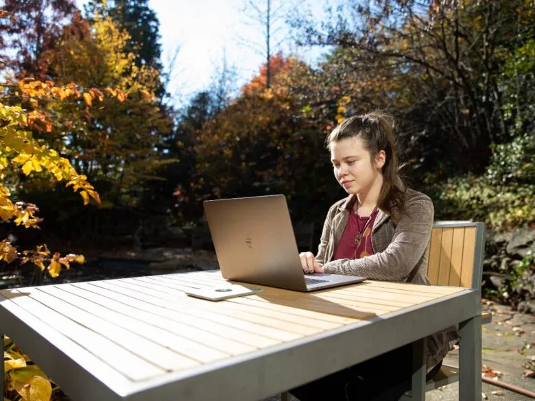 A student sitting outside during the day at a table, working on a laptop.