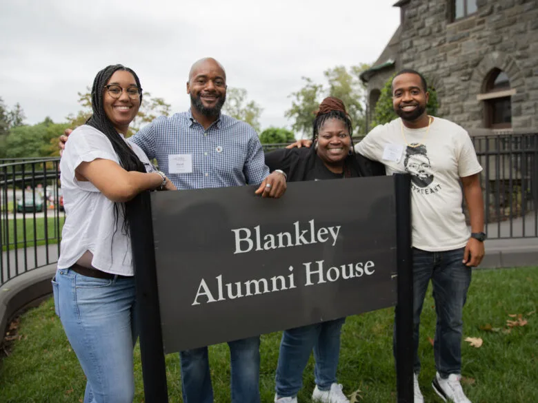 Alumni smiling at the camera and standing with the "Blankley Alumni House" sign.