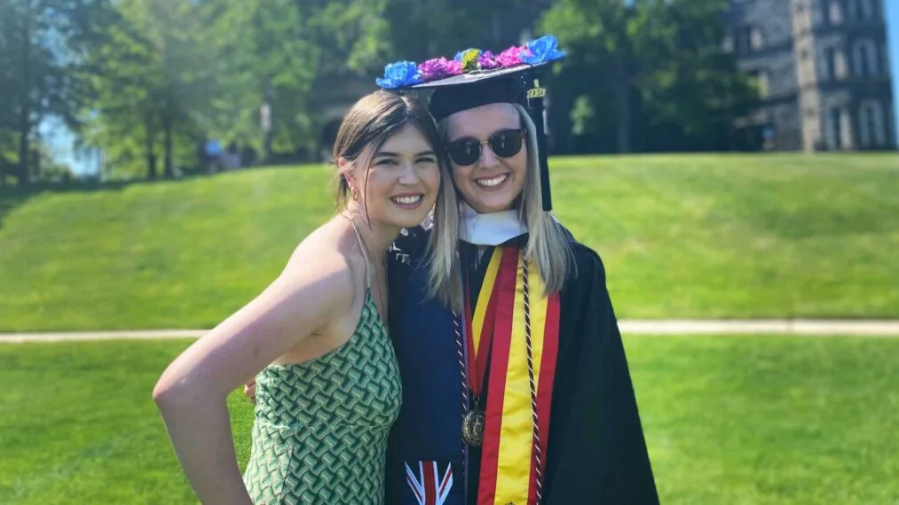 Graduate in cap and gown and friend smile at the camera while standing outside.