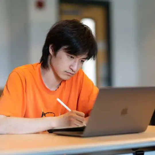 Student concentrating while looking at his laptop and taking notes at a desk.