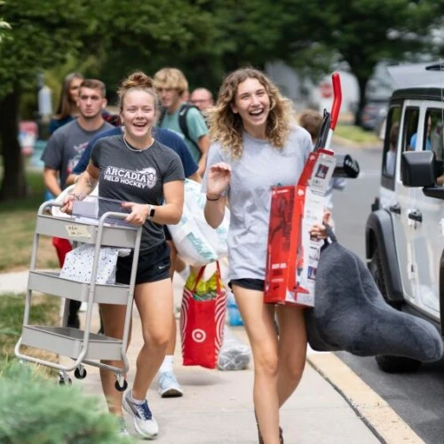 A group of students walking down a street carrying groceries and organizers and a pillow