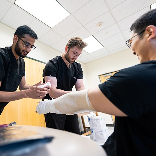 Three physician assistant students work on their medical skills gauzing an arm in a classroom setting.