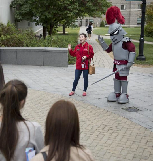 Archie stands sword in hand with a staff member in front of a crowd of undergraduate students on campus.